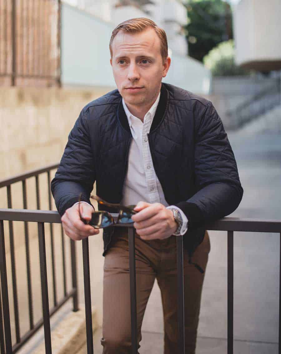 A man standing next to a fence wearing a bomber jacket and khaki pants