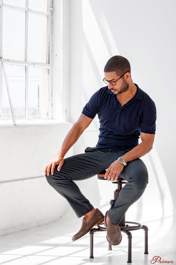 a man in glasses wearing a short sleeve henley and gray pants on a stool