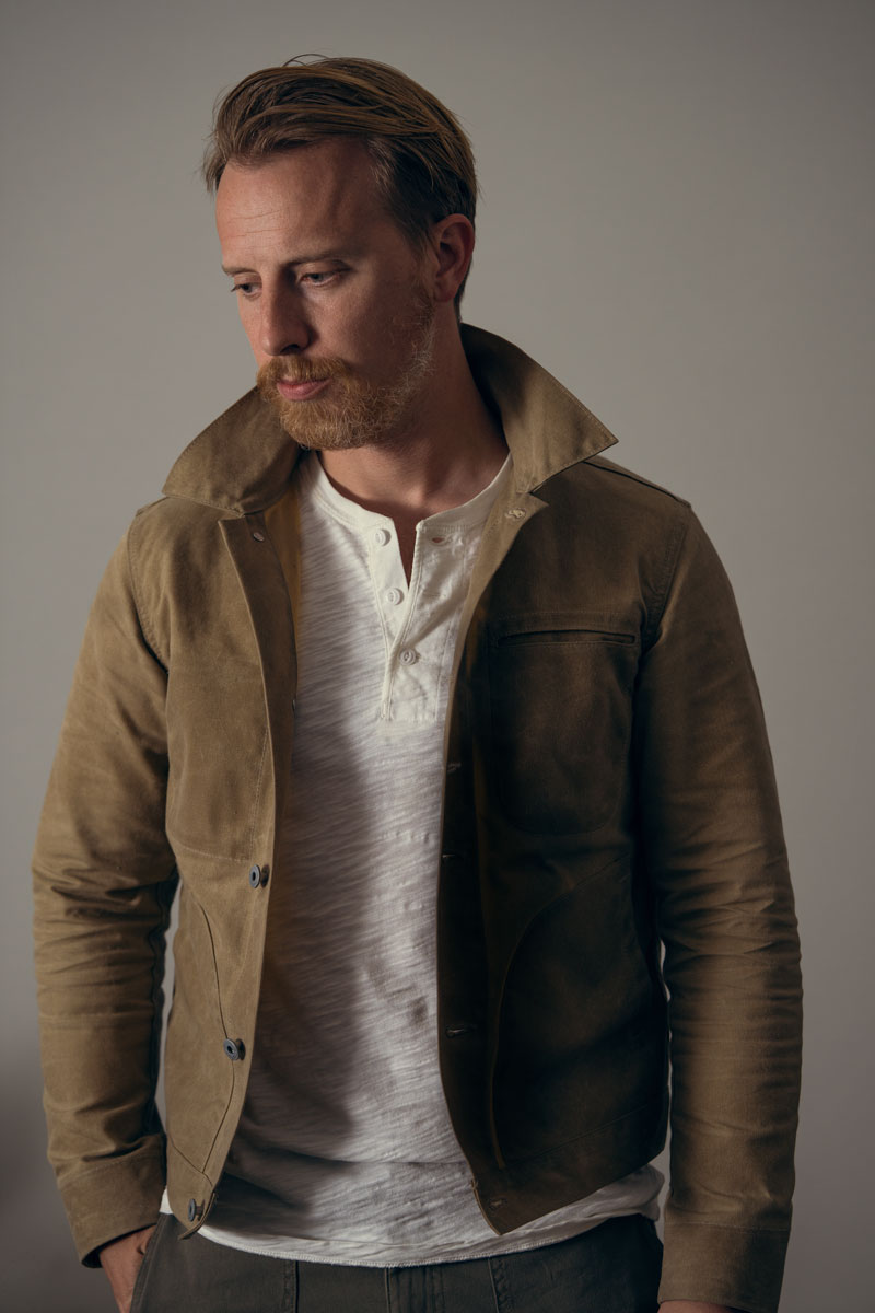 a man wearing a waxed canvas jacket over a shirt and jeans, with a relaxing dog in the rear compartment of a vehicle in the background