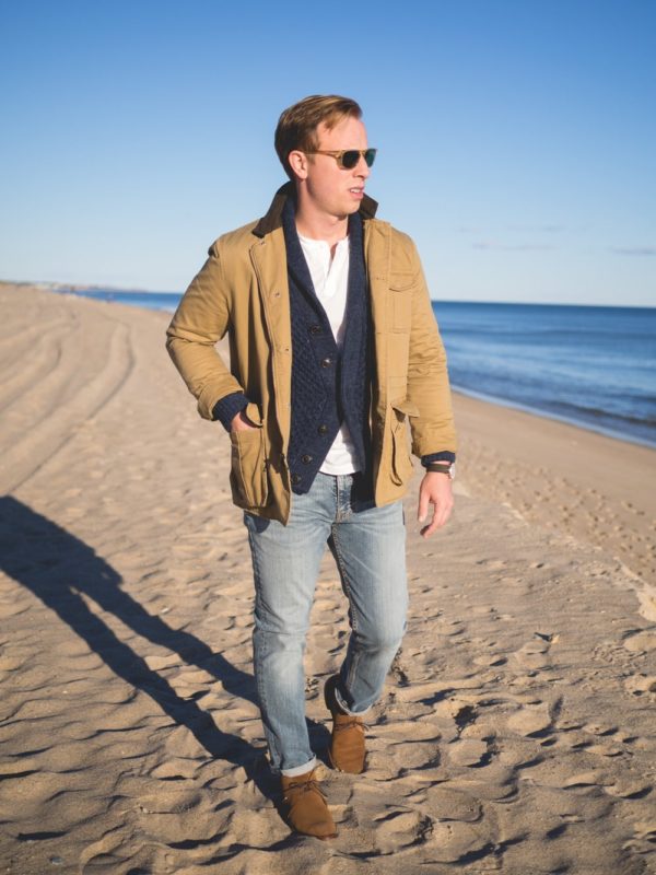 man wearing peter manning field coat with shawl collar cardigan, henley shirt, lightwash jeans, suede boots on a beach
