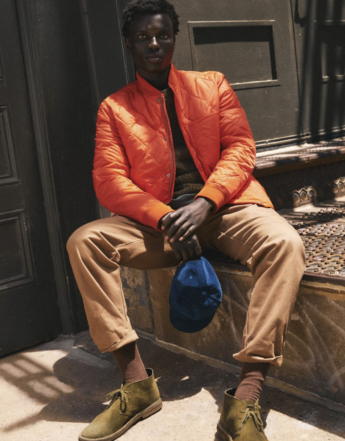 jcrew7 man sitting down wearing an orange jacket khaki pants and holding a blue hat