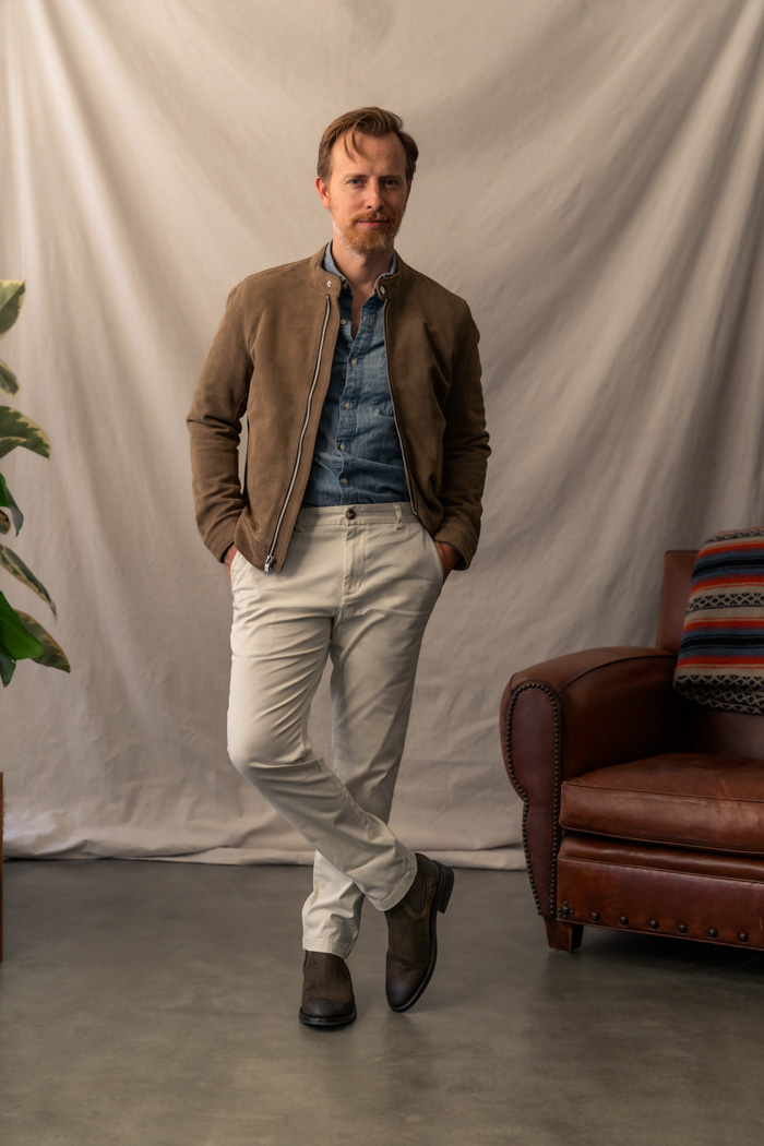 Full body photograph of a men's outfit with tan suede jacket, blue chambray shirt, white chino pants, and olive suede chelsea boots