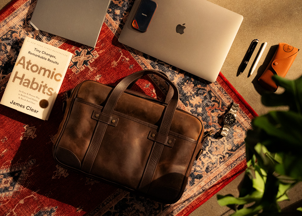 An overhead view of a scene showcasing a brown leather Thursday Boots Co. messenger bag, a copy of 'Atomic Habits' by James Clear, an Apple MacBook, a mobile phone, a pen, a leather pouch, and a wristwatch, all laid out on a richly patterned rug with a plant peeking into the frame.