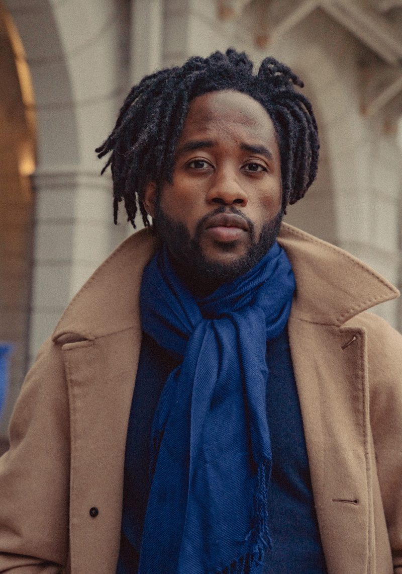 a man wearing a camel color topcoat and a vibrant blue scarf featuring an elegant knot with the end resting on top and in front of the rest of the scarf