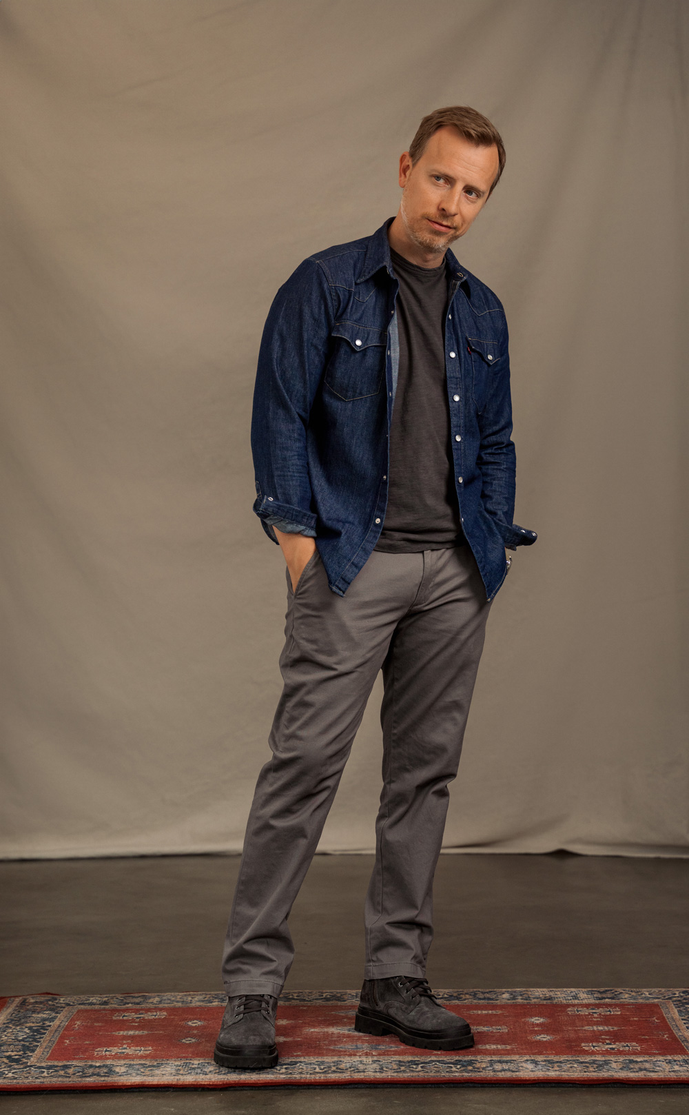 A man wears a dark grey t-shirt under an unbuttoned dark blue denim shirt and chest pockets over a black pocket t-shirt. His outfit is completed with grey trousers and dark grey suede boots. He has short blonde hair, and he poses with one hand in his pocket.