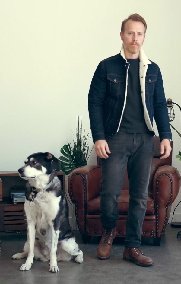 a man standing alongside a dog and wearing a sherpa lined jean jacket over a casual shirt black jeans and brown boot
