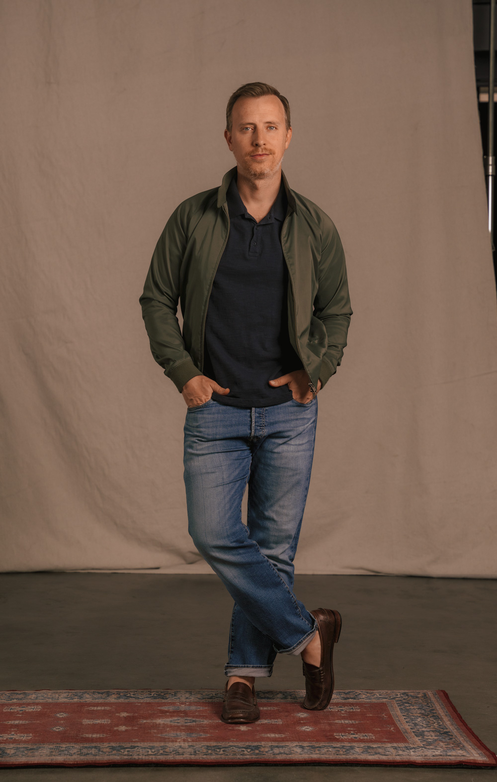 A man with short light brown hair and a light beard stands with his hands in his pockets. He is wearing a dark green jacket, a dark blue polo shirt, blue jeans, and brown loafers. The background is a plain beige curtain, and he stands on a red and blue patterned rug. One leg is crossed in front of the other.