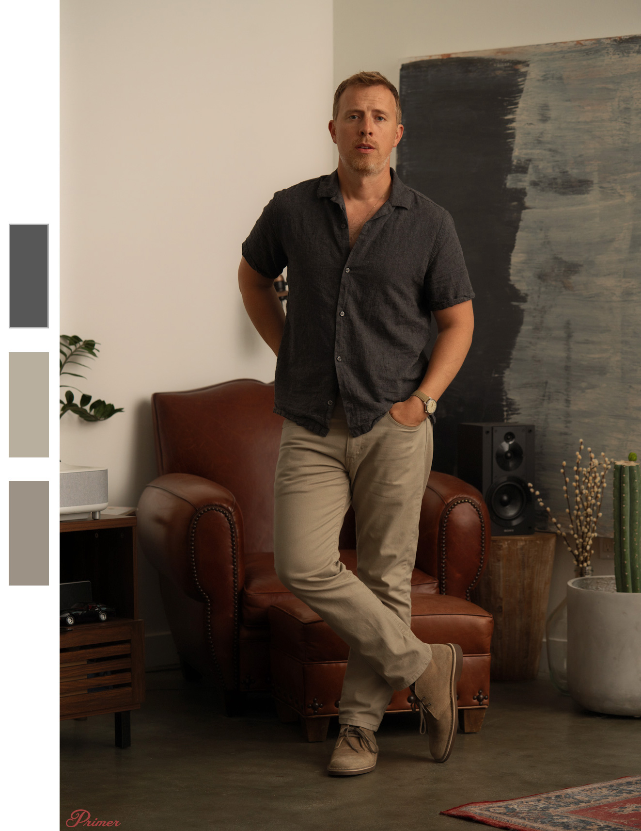A man stands casually with one leg crossed over the other in a modern, minimalist room. He is wearing a dark short-sleeve button-up shirt, beige pants, and light brown desert boots. Behind him is a leather armchair, a large abstract painting with dark tones, a small wooden table with speakers, and a potted cactus. The setting is well-lit with neutral tones, giving a calm and relaxed atmosphere.