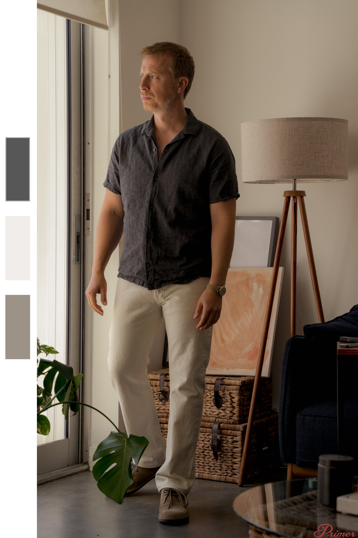 A man stands by a sliding glass door, looking out with a contemplative expression. He is dressed in a dark short-sleeve button-up shirt, off-white pants, and light brown desert boots. The room features a floor lamp with a beige shade on a wooden tripod base, wicker storage baskets, a framed abstract painting resting on the floor, and a large green monstera plant near the door. The overall aesthetic is warm and inviting, with natural light filtering into the space, highlighting the neutral and earthy tones of the decor.