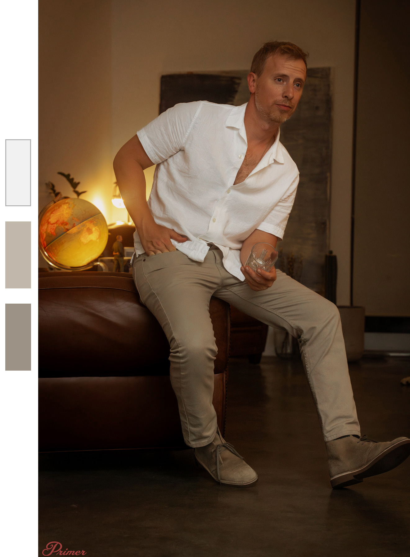 A man sits on the arm of a leather chair, wearing a white short-sleeve button-up shirt, beige pants, and light brown desert boots. He holds a glass tumbler in his right hand while resting his left hand on his hip. The room is warmly lit, with a glowing globe lamp and other small decor items on a nearby table. In the background, there is a large abstract painting, a potted cactus, and soft ambient lighting that enhances the relaxed and cozy atmosphere.