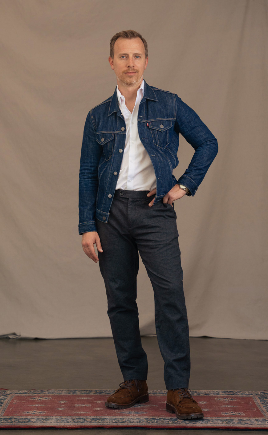 Andrew wearing a denim jacket over a white dress shirt with gray textured trousers with brown suede boots