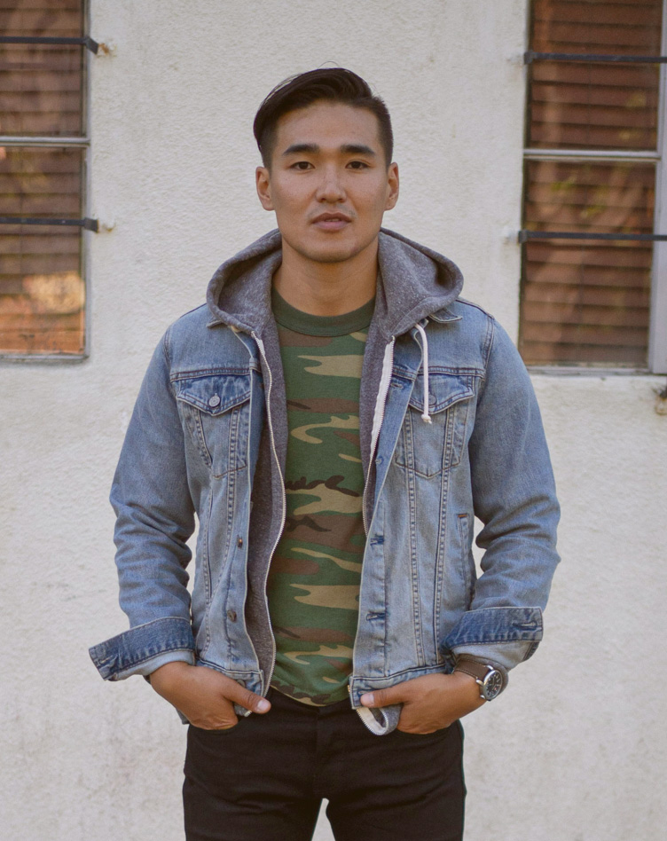 a man wearing a light wash jean jacket outfit over a camo pattern shirt black jeans and brown boots