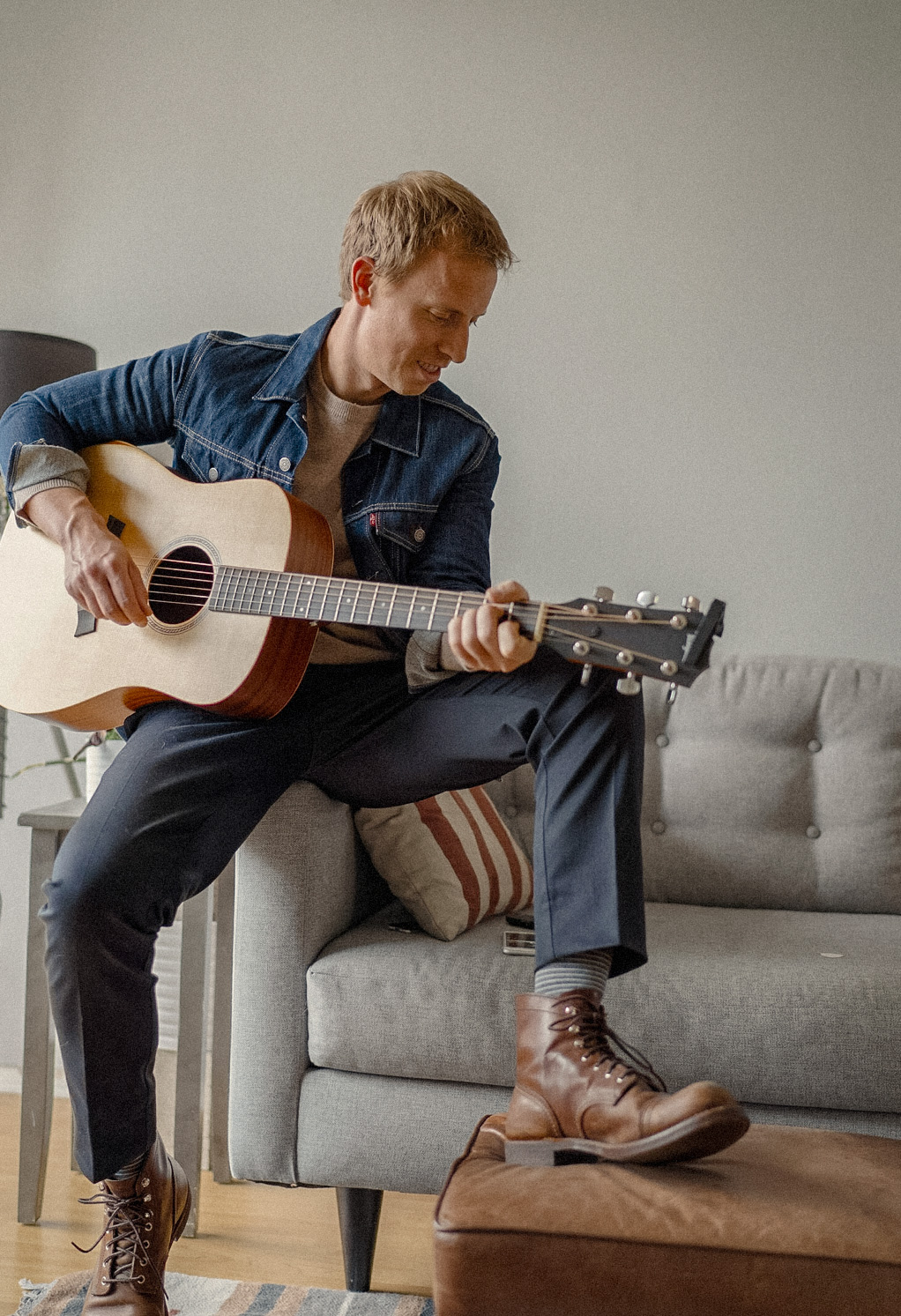 Andrew Snavely holding a guitar while waring a denim jacket over a tan colored sweater, black pant,s and brown Red Wing boots