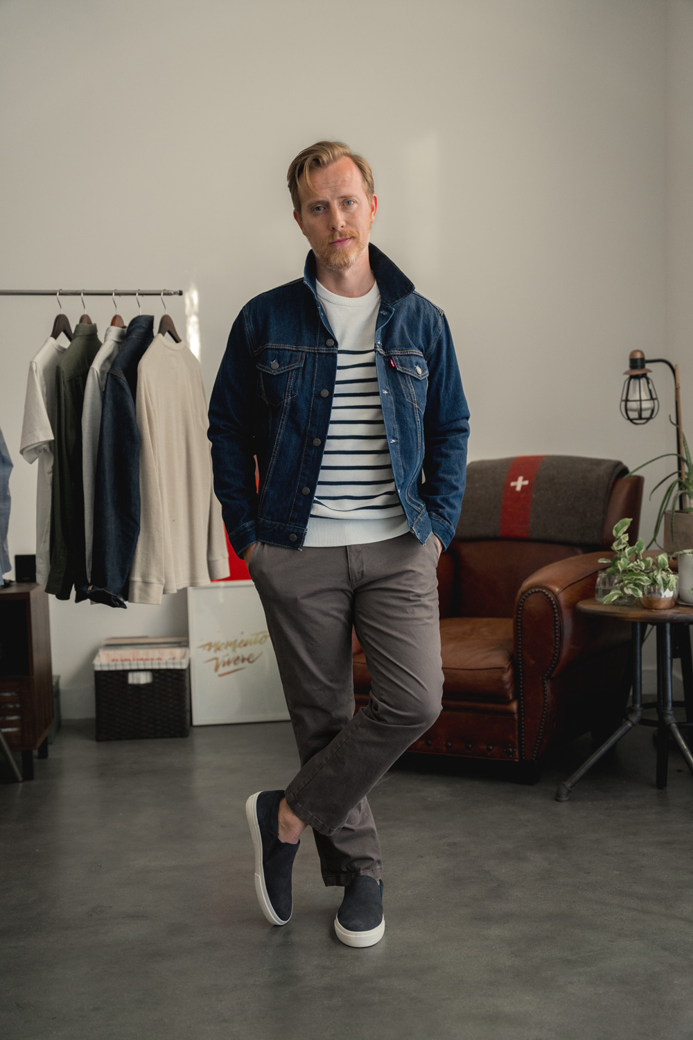 a man wearing a jean jacket over a striped shirt with slim fit pants and slip on shoes