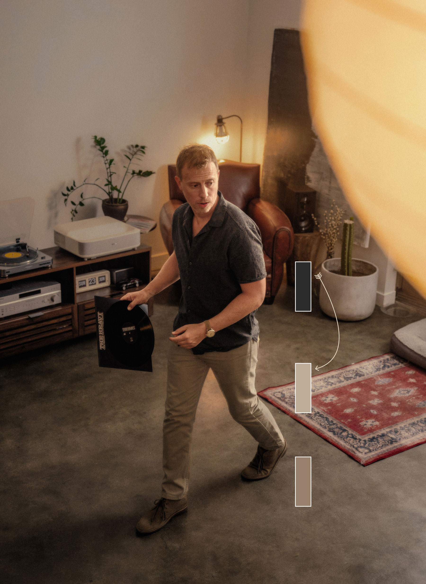 A man walks through a modern, minimalist living space while holding a vinyl record. He is dressed in a dark short-sleeve button-up shirt, beige pants, and light brown desert boots. The room includes a wooden console with a record player and audio equipment, a leather armchair, a small table with a potted plant, and a large abstract painting. The lighting is warm, with a lit lamp near the armchair and a soft, glowing light fixture partially visible in the foreground. A red and blue patterned rug and a large potted cactus add decorative touches to the space.