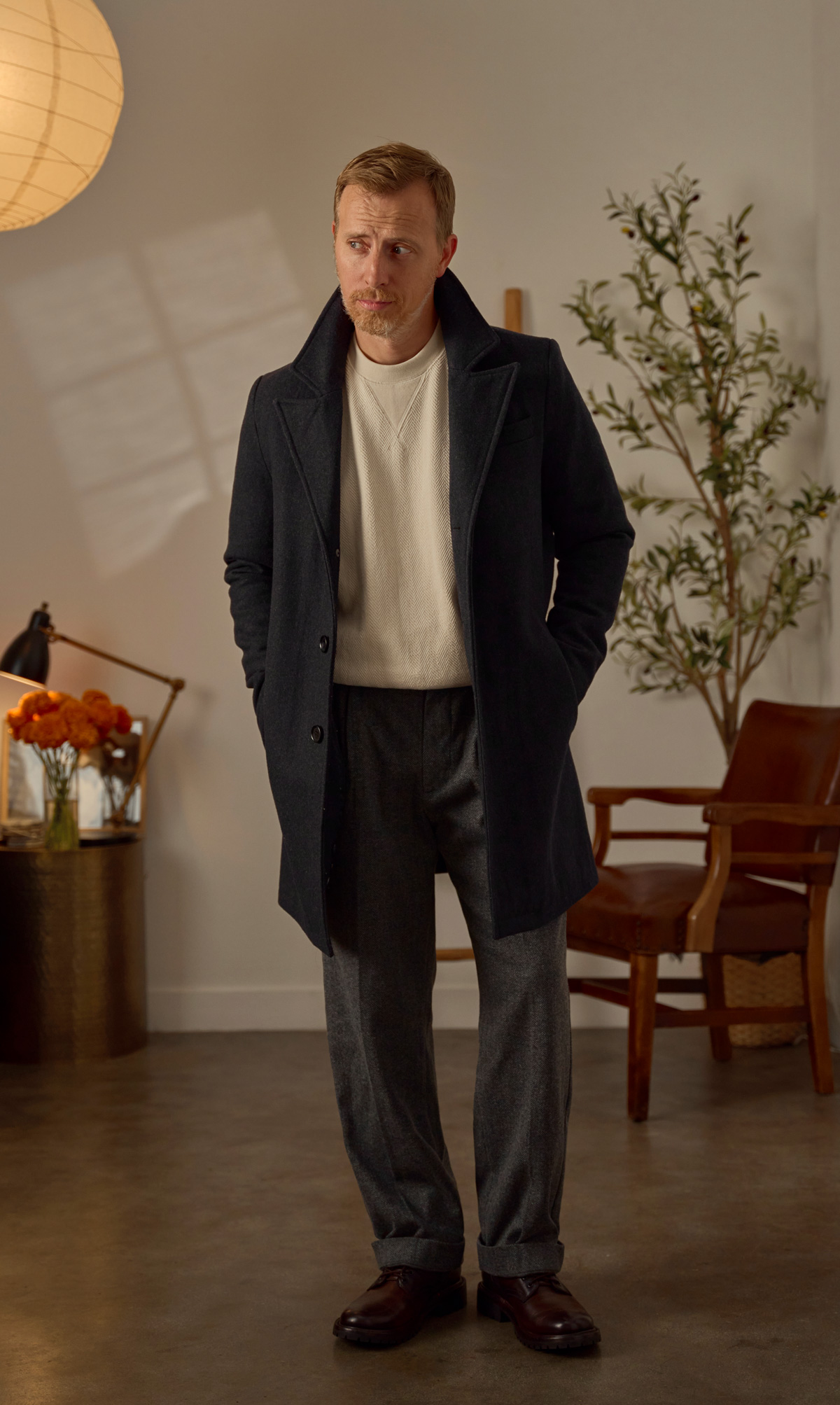 Andrew wearing a holiday outfit for men consisting of a dark gray topcoat over a beige herringbone sweater with gray fuller cut trousers and brown boots