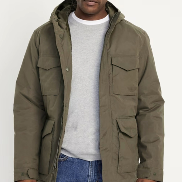 a man wearing a utility style jacket over a crew neck sweater and denim jeans