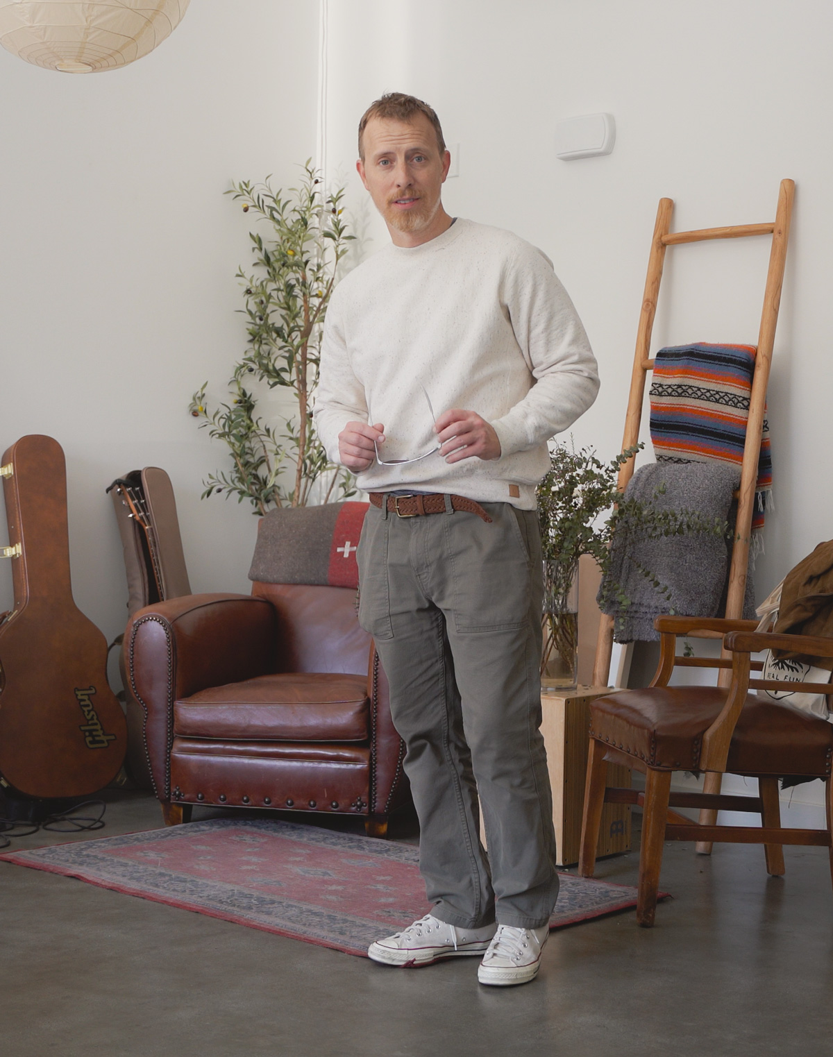 a man with short, light brown hair and a beard standing in a well-lit living space. He wears an outfit similar to the one in the collage: a cream-colored crewneck sweatshirt, olive green utility pants, a brown braided leather belt, and off-white Converse sneakers. He holds a pair of eyeglasses in both hands and looks directly at the camera with a relaxed expression. The setting includes a brown leather armchair with a red and gray patterned cushion, a wooden ladder leaning against the wall holding a striped blanket, a potted plant, a wooden chair, and a guitar case labeled “Gibson.” A red and gray rug lies on the polished concrete floor. The room has white walls and natural light coming from an unseen window.