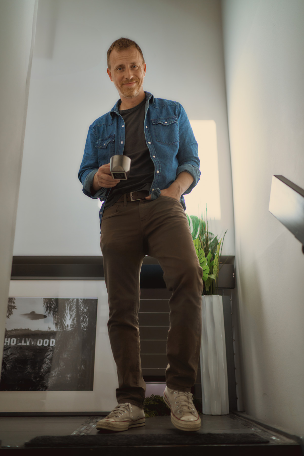a man wearing a chambray shirt over a casual crew neck shirt, chino pants, and lace up sneakers