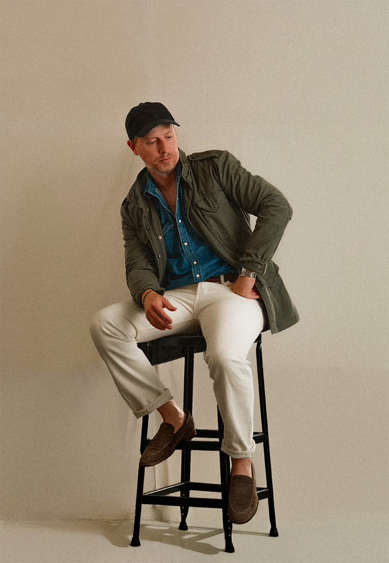 A man sitting casually on a tall stool against a neutral background. He is wearing a black baseball cap, an olive-green jacket, a denim shirt, white pants, and brown loafers. He has one hand in his pocket and is looking slightly downward.