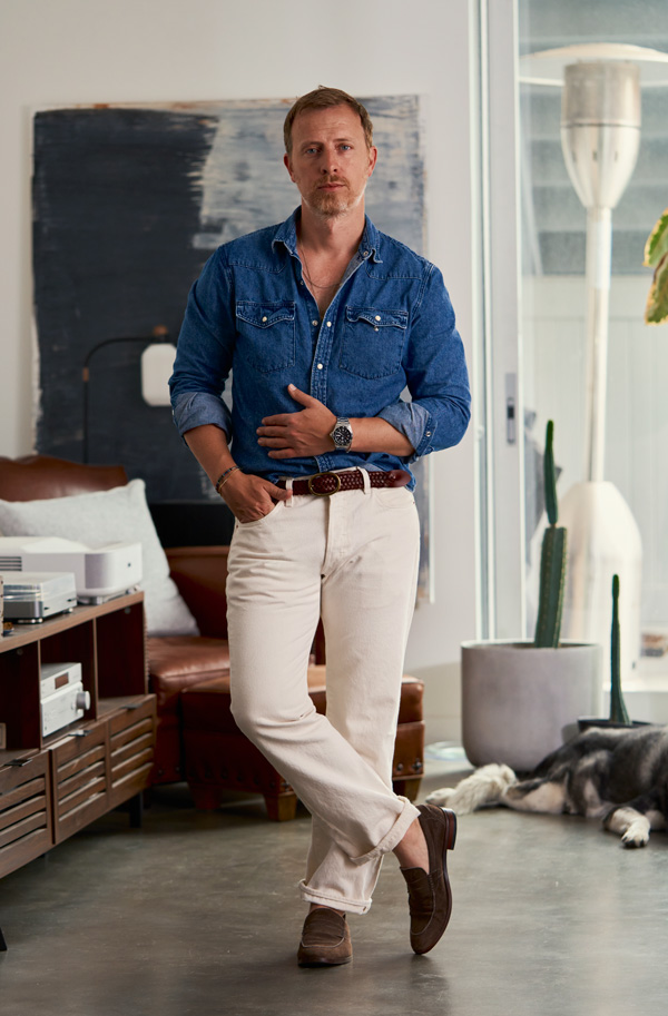 A man sitting casually on a tall stool against a neutral background. He is wearing a black baseball cap, an olive-green jacket, a denim shirt, white pants, and brown loafers. He has one hand in his pocket and is looking slightly downward.