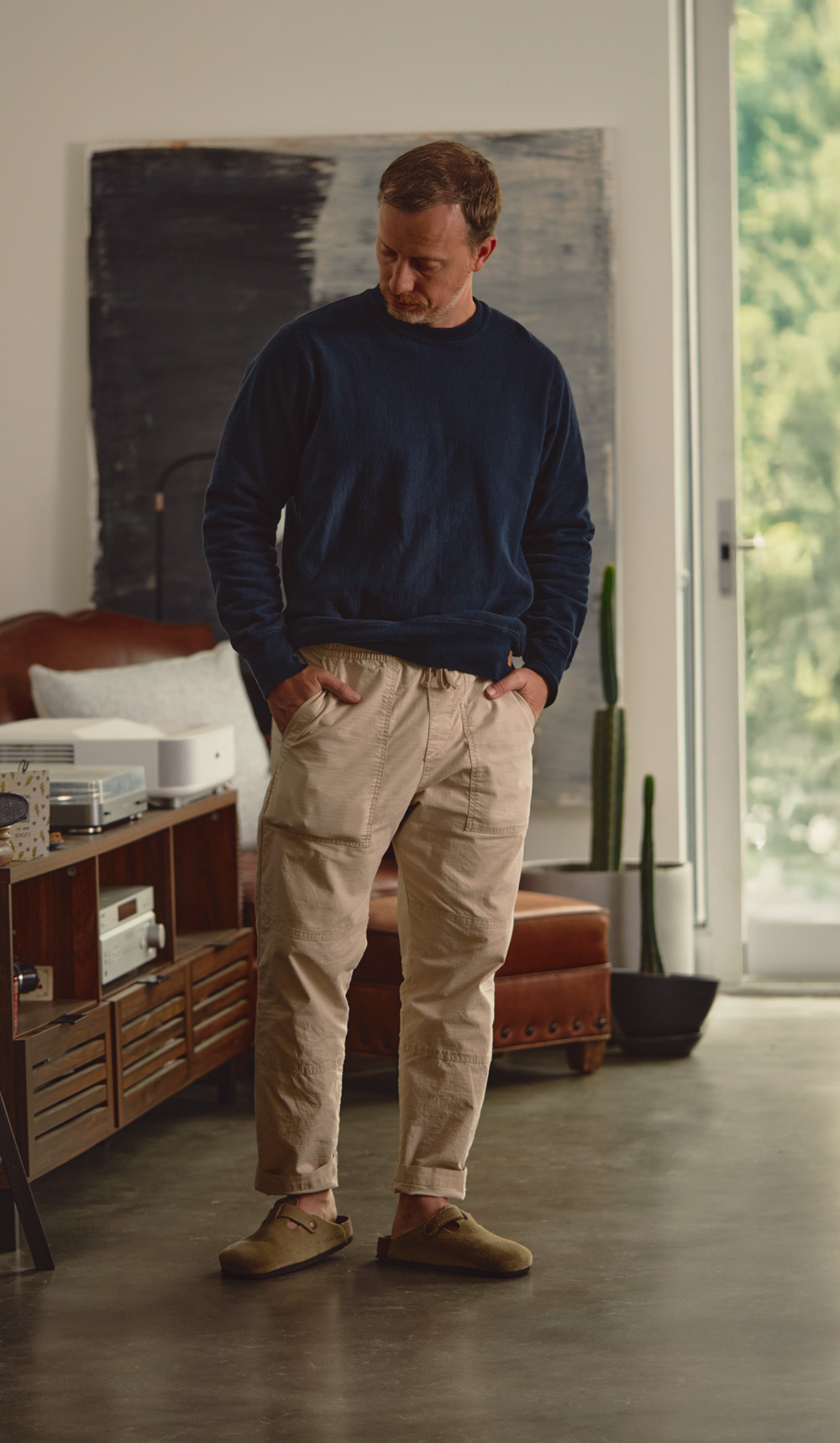 Man standing indoors with his hands in the pockets of beige drawstring pants, wearing a dark blue long-sleeve sweatshirt and taupe suede slip-on shoes. He is looking down slightly, with a relaxed posture. Behind him is a brown leather chair with a pillow, a wooden cabinet with audio equipment, a tall cactus in a planter, and a large abstract painting on the wall. Natural light comes through a glass door on the right.