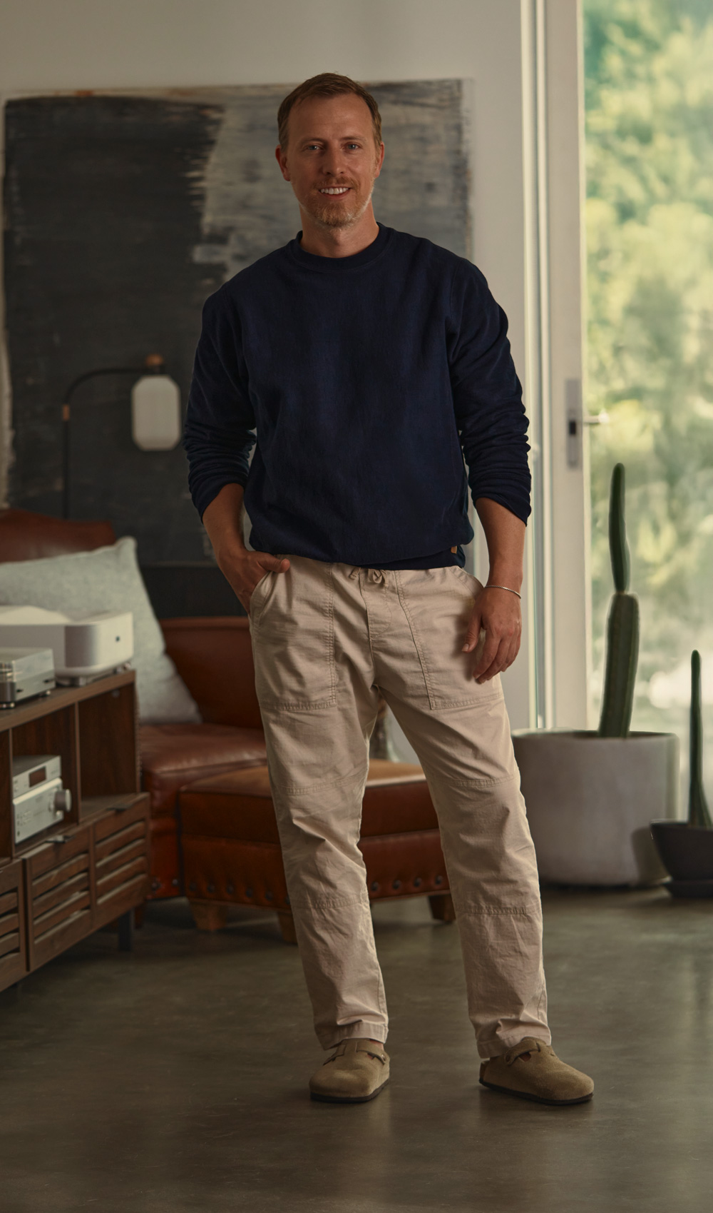 Andrew standing indoors with his hands in the pockets of beige drawstring pants, wearing a dark blue long-sleeve sweatshirt and taupe suede slip-on shoes. He is looking down slightly, with a relaxed posture. Behind him is a brown leather chair with a pillow, a wooden cabinet with audio equipment, a tall cactus in a planter, and a large abstract painting on the wall. Natural light comes through a glass door on the right.
