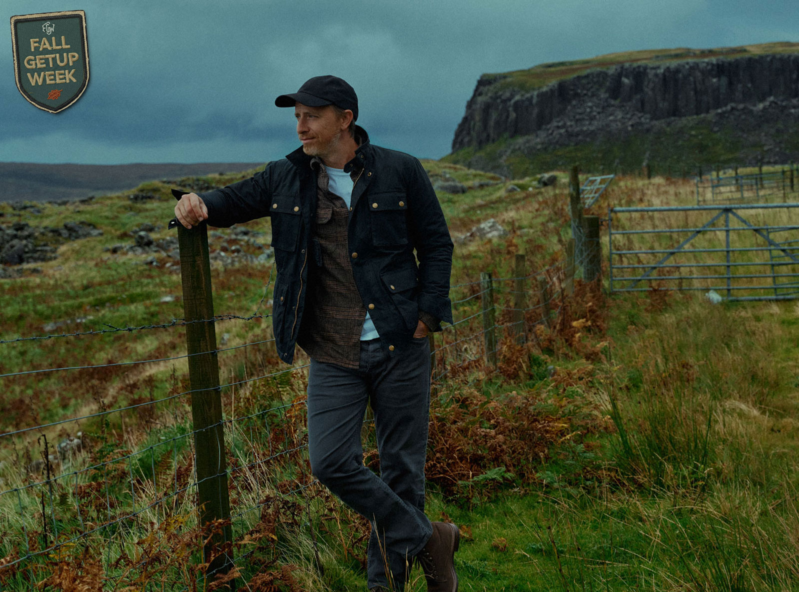 A man wearing a fall outfit stands outdoors leaning against a wooden fence post in a rugged rural landscape with grassy fields, patches of ferns, and rocky terrain. He is wearing a dark jacket, gray pants, brown boots, and a black cap, looking off to the side with one hand in his pocket. A metal gate and steep cliffs are visible in the background under a cloudy, moody sky. a badge reads fall getup week in the corner