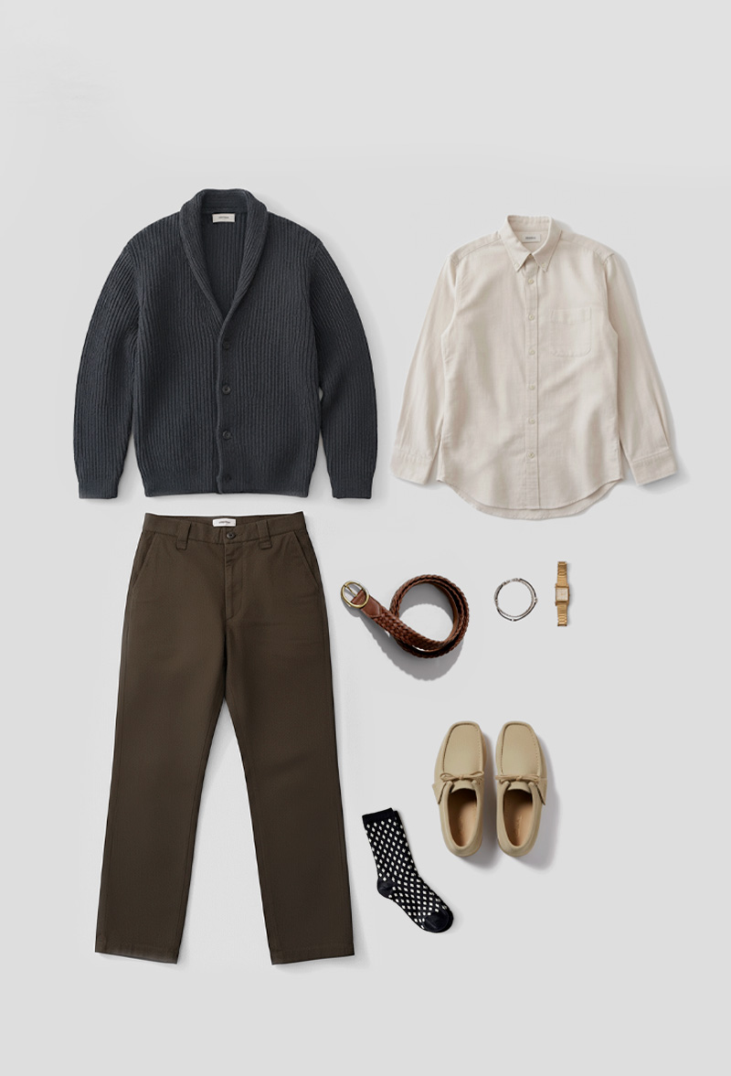 fall-outfit-classic-fit_2 A flat lay of a smart casual men's outfit arranged on a light grey background. The ensemble includes a dark grey ribbed shawl-collar cardigan, an off-white button-down shirt, and a pair of olive green chinos. Accessories include a braided brown leather belt, a silver bracelet, a gold-toned wristwatch, and a pair of black socks with white polka dots. The shoes are light beige suede moccasin-style loafers.