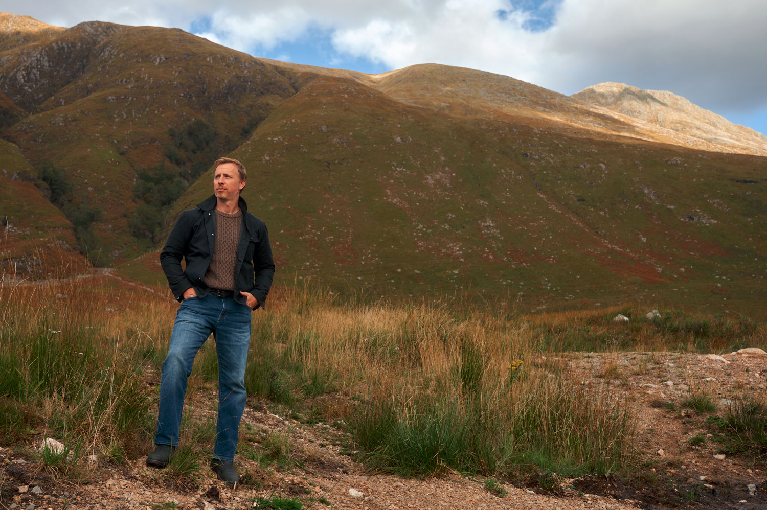 mens fall outfit idea featuring black waxed canvas jacket with brown sweater, jeans, and black boots