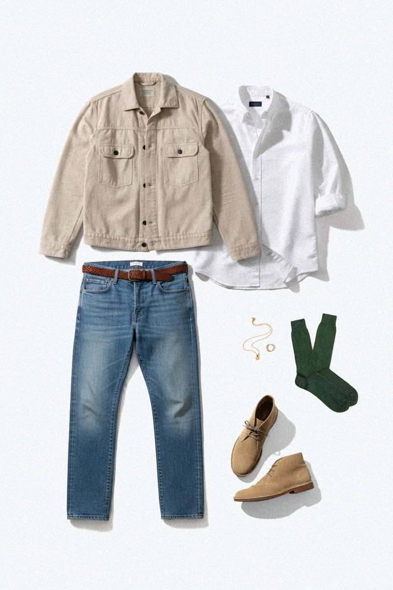 lightweight-fall-outfit_1-flat A flat lay of a lightweight men's casual fall outfit arranged on a white surface. The items include a beige shirt-jacket, a white button-down shirt, blue jeans with a brown braided belt, green socks, tan suede chukka boots, a gold necklace, and a gold ring.