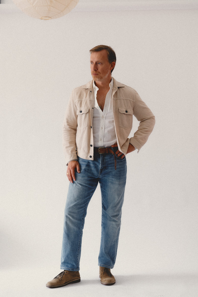 lightweight-fall-outfit_1 A man stands in a full-body shot against a plain off-white background. He wears a beige shirt-jacket with black buttons, a white button-down shirt, blue jeans with a brown braided belt, and tan suede chukka boots. One hand is in his pocket and the other rests at his side.
