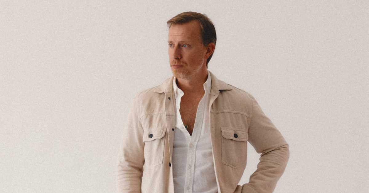 A man in a light fall outfit for warm weather poses indoors against a plain off-white background. He is wearing a beige shirt-jacket with black buttons over a partially unbuttoned white shirt with a necklace