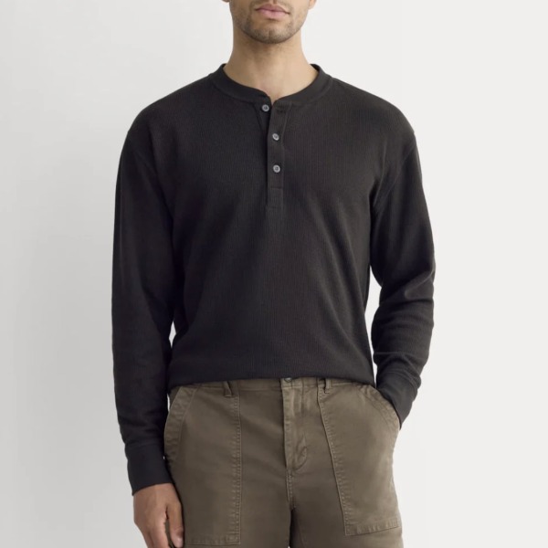 A man wearing a black waffle knit thermal henley shirt with three buttons, paired with light-colored pants, standing against a plain white background.