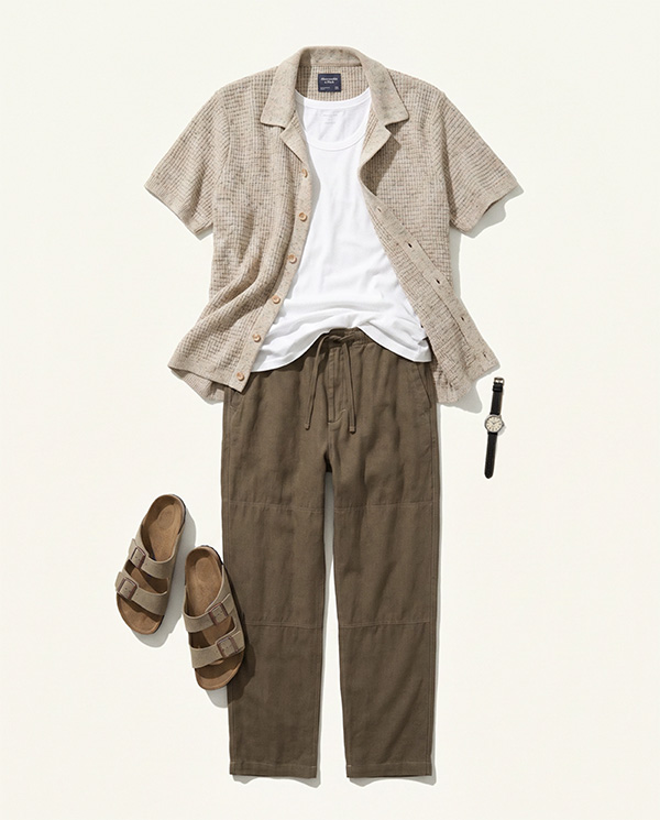 Flat lay outfit featuring a beige short-sleeve textured button-up shirt worn open over a white crew-neck T-shirt, paired with loose brown drawstring pants. Brown double-strap sandals and a black wristwatch are arranged beside the clothing on a light neutral background.